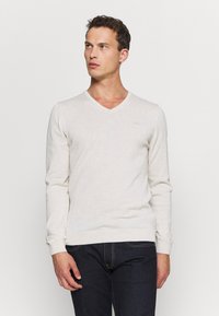 Mann in hellbeigem V-Ausschnitt-Pullover und dunklen Jeans, der vor einem schlichten weißen Hintergrund steht und zur Seite schaut.
