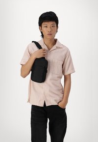 Bolso bandolera negro con compartimentos con cremallera. El modelo lleva una camisa de manga corta a rayas rosas y pantalones negros, sosteniendo el bolso con una mano.
