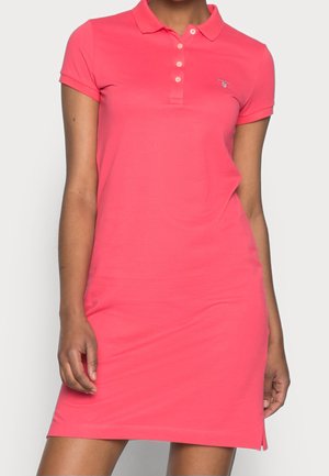 Kobieta w jasnoróżowej sukience koszulowej z krótkim rękawem, z kołnierzykiem zapinanym na guziki i małym logo na lewej piersi.