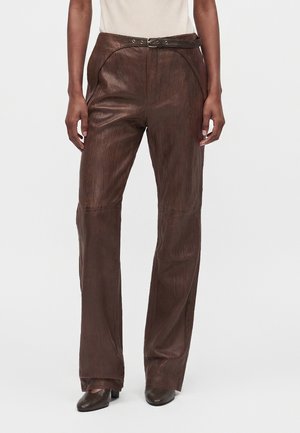 Personne portant un pantalon en cuir marron foncé taille haute avec une ceinture assortie et des chaussures marron à bout pointu sur fond blanc.