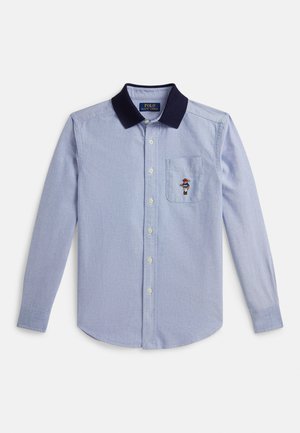 Svetlo modra majica z dolgimi rokavi, temno mornarsko modrim ovratnikom, gumbi spredaj, prsnim žepom in majhnim vezenim medvedom, ki na žepu drži palico.