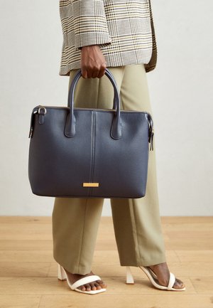 Personne portant un pantalon beige et une veste à carreaux tient un grand sac à main bleu marine, debout sur un sol en bois avec des sandales blanches à talons.