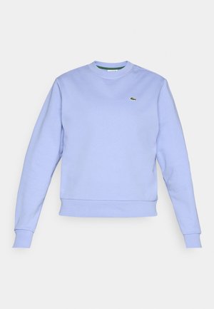 Personne en sweat-shirt bleu clair, jupe de tennis blanche, baskets blanches tenant une raquette de tennis et une balle de tennis verte, debout devant un fond uni.