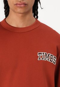 Rust-kleurig sweatshirt met geribbelde kraag, met het woord "TIMBS" in witte geborduurde letters op de linkerborst. Voelt soepel aan.