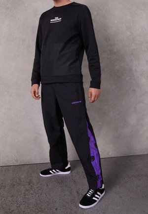 Czarna koszula z długim rękawem z logo, w zestawieniu z czarnymi spodniami z fioletowym paskiem po boku i wzorzystymi akcentami. Czarne sneakersy.