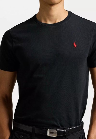 T-shirt nera a girocollo realizzata in tessuto a costine. Presenta un piccolo logo ricamato rosso sul petto sinistro. Indossata con una cintura nera.
