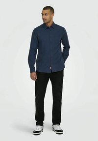 Camicia blu navy a maniche lunghe con bottoni e tasca sul petto, abbinata a pantaloni neri e sneakers bianche. Il materiale appare intrecciato con una texture liscia.