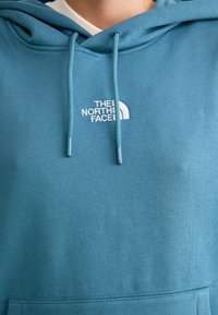 Sweat à capuche turquoise en mélange de coton, avec un logo brodé blanc et des cordons, mettant en avant une surface texturée.