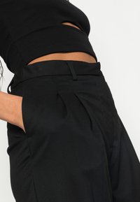 Svarta plisserade byxor med hög midja och sidofickor, tillverkade av ett mjukt tyg med en subtil textur. Framträdande med en åtsittande midjeband.