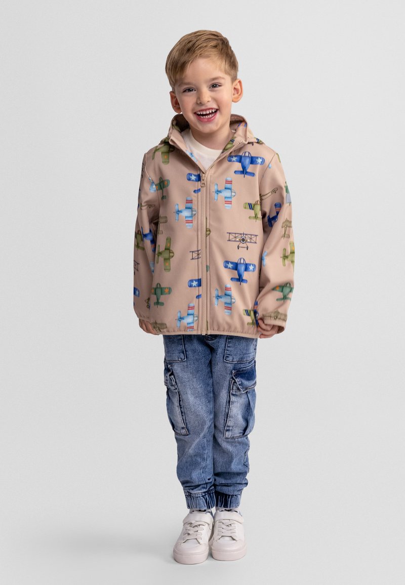 Jeune garçon souriant portant une veste beige avec des imprimés d'avions colorés, un jean cargo bleu et des baskets blanches, debout devant un fond uni.