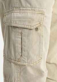 Pantalons cargo beige avec deux poches latérales, des coutures renforcées et un tissu doux et texturé. Le matériau semble durable et léger.