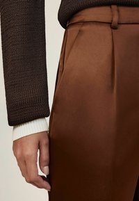 Barna selyem nadrág oldalsó redőkkel, texturált sötétbarna kötött pulóverrel és fehér bordázott mandzsettával párosítva. Simogató anyag, testhezálló szabás.