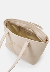 Ljusbeige läder toteväska med långa remmar, texturerad yta och ett inre fack. Utrustad med guldfärgad metall och präglad text.