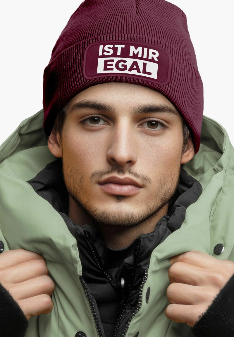 Maroon knit beanie with white "IST MIR EGAL" text. Green puffer jacket with black accents. Hands gripping jacket, showing textures and layers.