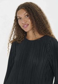 Donna con lunghi capelli ricci e castani, che indossa un top lungo a maniche nere texture, che sorride leggermente su uno sfondo bianco neutro.