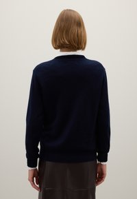 Marineblauwe wollen trui met een ronde hals, geribbelde manchetten en zoom, en een gladde textuur. Duidelijke witte kraag van een overhemd eronder zichtbaar.