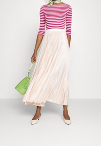 Person, die ein pink-weiß gestreiftes Oberteil, einen hellrosa gefalteten Maxirock, beige Absatzschuhe trägt und eine kleine grüne Handtasche hält.