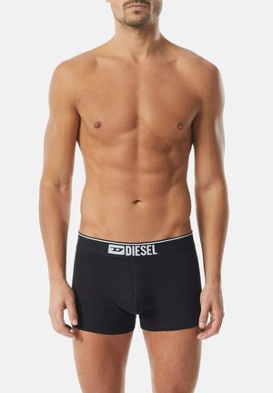 Oberkörperfreier Mann trägt schwarze Diesel-Boxershorts mit Logobund und steht vor einem schlichten hellen Hintergrund.