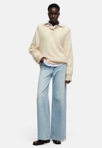 Maglione di lana beige chiaro con collo e due bottoni. Abbinato a jeans a gamba larga azzurri e calzature marrone scuro. Look semplice e strutturato.