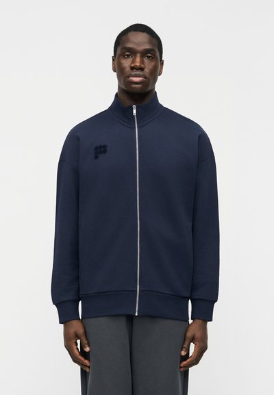 Homme portant une veste zippée bleu marine avec un petit logo brodé sur le côté gauche de la poitrine, debout devant un fond clair uni.