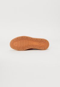 Suela de zapato con una banda de rodadura de goma marrón que presenta patrones geométricos y rectangulares sobre un fondo blanco.