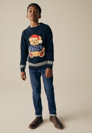 Marinblå stickad tröja med ett nallebjörnsdesign, röd tomteluva och snöflinge mönster; kombinerad med blå jeans och bruna ankelstövlar.