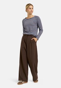 Frau steht mit den Händen in den Taschen, trägt ein blau- und braun gestreiftes Langarmshirt, weite braune Hose und braune Sneakers mit weißen Streifen.