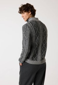Maglione grigio lavorato a maglia con un motivo a treccia testurizzato e colletto alto, abbinato a pantaloni grigi scuri. La vista dal retro mostra dettagli intricati.