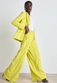 Geel op maat gemaakte blazer met bijpassende wijde broek, gemaakt van gladde stof. Witte fitted top eronder, aangevuld met zwarte hoge hakken.