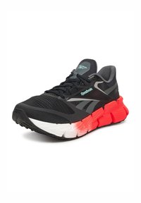 Scarpa sportiva nera con tomaia in mesh, dettagli grigi e una suola spessa multicolore con sezioni rosse e bianche. Logo Reebok visibile.