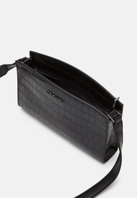 Czarna torba ze skóry tłoczonej na wzór krokodyla, o płaskim kształcie, z zapinanym na zamek komorą i regulowanym paskiem dla różnorodnych sposobów noszenia.