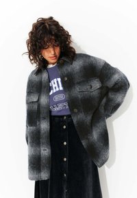 Veste en laine grise et noire à carreaux surdimensionnée avec deux poches sur la poitrine, des fermetures à bouton et une texture douce, superposée sur un sweat-shirt violet.