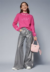 Pink knitted sweater with heart patterns, silver flared pants, and a small pink handbag with a fruit design. White sneakers complete the look.
