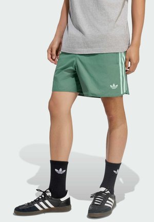 Person, die grüne Adidas-Shorts, schwarze Adidas-Socken, schwarz-weiße Adidas-Sneaker und ein graues T-Shirt trägt, gezeigt vom Taillenbereich bis zu den Füßen.
