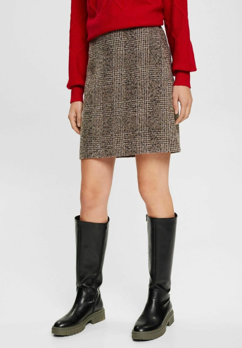 Femme portant une mini-jupe à carreaux marron, un pull rouge et des bottes noires montantes, debout devant un fond clair.