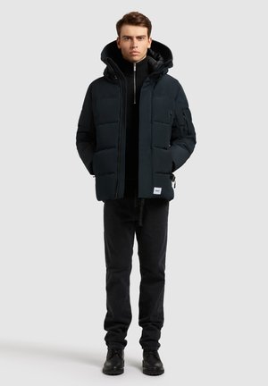 Chaqueta acolchada oscura con capucha, que cuenta con bolsillos laterales con cremallera y un cierre frontal con cremallera. Combinada con jeans negros y botas negras.