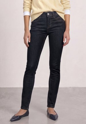 Persona con vaqueros ajustados oscuros, suéter beige con mangas remangadas y zapatos planos oscuros con punta, de pie sobre un suelo liso.