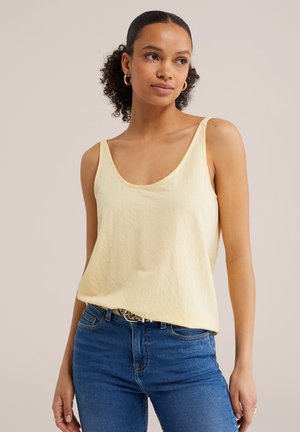 Femeie cu păr creț, purtând cercei hoop aurii, un top fără mâneci de culoare galben deschis, blugi albaștri și o curea, pe un fundal simplu.