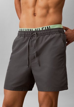 Mandetorso iført mørkegrå badeshorts med elastisk talje og snor, delvist synlig grøn Calvin Klein undertøjslinning.