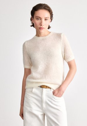 Krátky rukáv sveter z jemného krémového priadze. Má okrúhly výstrih a rebrovaný spodek. Nosený s bielymi nohavicami a svetlým opaskom.