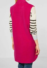 Rosa ärmlös väst med hög krage, tillverkad av mjukt material. Bärs över en randig långärmad skjorta med ribbade manschetter.