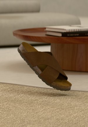 Smeđa sandala s križnim trakama i plutenim đonom lebdi iznad bež tepiha, okruglog drvenog stolića za kavu i neutralnog kauča u pozadini.