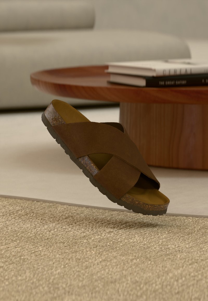 Καφέ σανδάλι με λουριά που διασταυρώνονται και σόλα από φελλό που αιωρείται πάνω από μπεζ χαλί, στρογγυλό ξύλινο τραπεζάκι καφέ και ουδέτερο καναπέ στο φόντο.