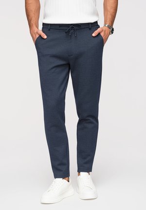 Homme portant un pantalon slim bleu marine avec taille à cordon et des baskets blanches, mains dans les poches, sur fond uni.