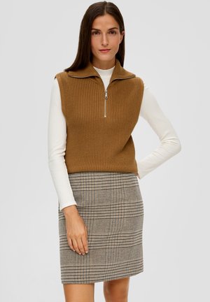 Bruine gebreide mouwloze trui met een hoge kraag en ritssluiting, gecombineerd met een grijze geruite kokerrok. Witte lange mouwen top eronder.