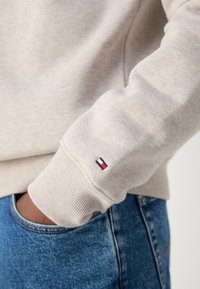 Lys grå sweatshirt med ribstrikkede manchetter, prydet med en lille logo-patch i blå, rød og hvid på ærmet. Båret sammen med blå denimjeans.