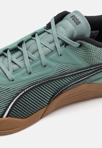 Zapatilla deportiva de malla verde con acentos negros, superficie texturizada y suela de goma. Presenta cordones finos y un diseño de parte superior que ofrece soporte.