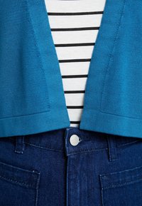 Turquoise knit cardigan over a white and black striped shirt, paired with dark blue denim jeans. Features a silver button on the waistband.