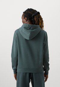 Tamsiai žalia su kapuca džemperis pagamintas iš minkštos, tekstūruotos medžiagos. Turi įprastą kirpimą ir ruoželinės rankogalius bei apačią. Minimalistinis dizainas.