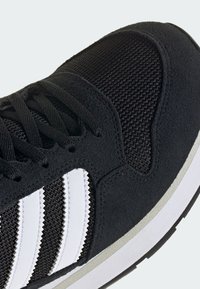Chaussure de sport noire avec trois bandes blanches sur le côté, lacets noirs, dessus en mesh, et détail de semelle blanc et gris.